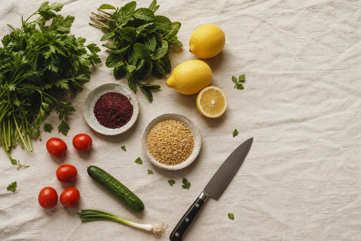 Salad ingredients laid out on linen: bunches of parsley and mint, lemons, small tomatoes, a cucumber, a spring onion and dishes of sumac and cracked bulgur.